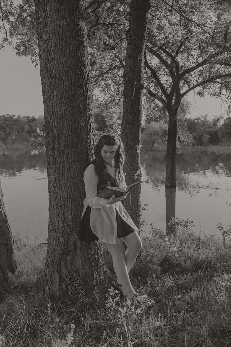 Woman sitting against a tree reading a book outdoors in black and white